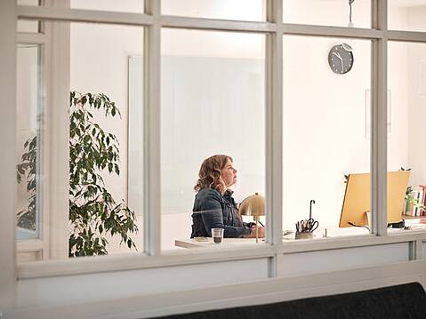 Bild von Lexware: Sicht von außen durchs Fenster auf eine Unternehmerin am Schreibtisch vorm Bildschirm